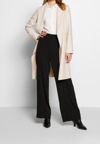 Abrigo largo beige con solapas anchas, combinado con una camisa blanca y pantalones de pierna ancha negros. Botines negros de punta completan el conjunto.