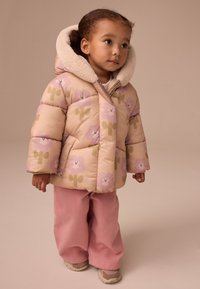 Gepolsterter, beiger Mantel mit rosa Blumenmustern, Kapuze mit Kunstpelz gefüttert; kombiniert mit rosa Hosen und beigen Sneakers. Glatte Textur, lässige Passform.