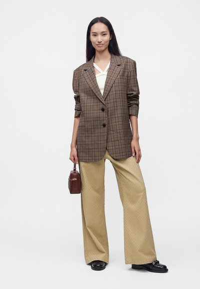 Femme debout portant un blazer à carreaux marron, un haut crème, un pantalon beige à jambes larges avec de petits pois, des chaussures noires et tenant un petit sac à main marron.