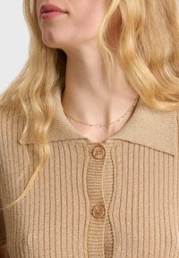 Haut beige à rayures avec boutons, fabriqué dans un matériau texturé, comportant un col classique et deux boutons ronds à l'avant. Détail de subtil éclat.