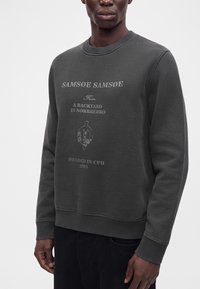 Homme portant un sweat-shirt gris foncé avec le texte "SAMSOE SAMSOE From A BACKYARD IN NØRREBRO FOUNDED IN CPH 1993" et un motif floral.