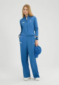 Chándal atlético azul con chaqueta de cremallera y pantalones de pierna ancha. Confeccionado en tejido suave, presenta acentos de logo. Incluye una gorra azul a juego.