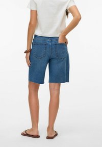 Shorts de mezclilla azul con dobladillo deshilachado, corte de tiro medio y diseño de cinco bolsillos; combinados con una blusa blanca y chanclas marrones.