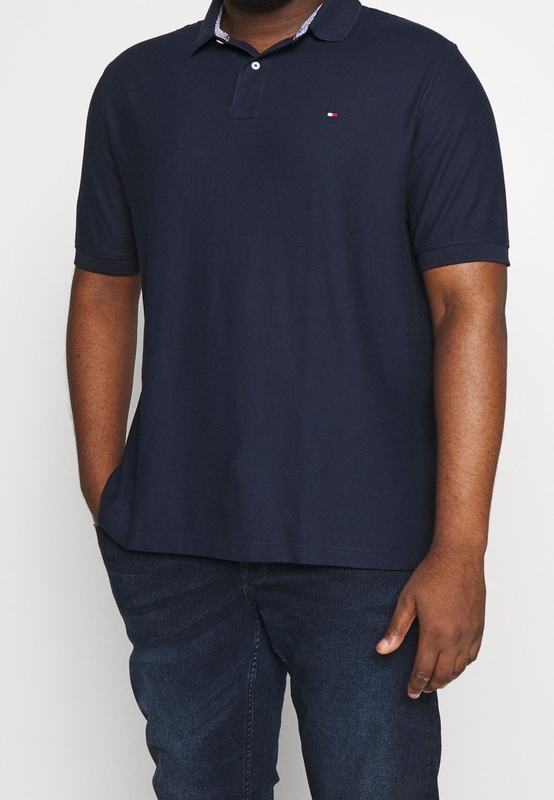 Homme portant un polo bleu marine à manches courtes et un jean bleu foncé, debout avec une main dans la poche sur un fond uni.