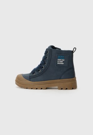 Botines de color azul marino hechos de material sintético, con suela de goma marrón, puntera redonda, seis ojetes metálicos y un parche con texto.