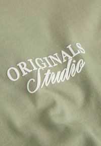 Λευκό κεντημένο κείμενο "ORIGINALS Studio" σε υφή υφάσματος ανοιχτού ελιάς με ελαφρές ορατές πτυχές υφάσματος.