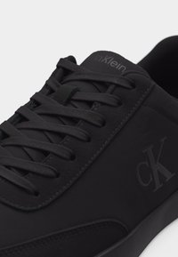 Μαύρο αθλητικό παπούτσι Calvin Klein με μαύρα κορδόνια, με το εμπορικό σήμα "ck" στο πλάι και ραφές γύρω από τις τρύπες των κορδονιών και τη σόλα.