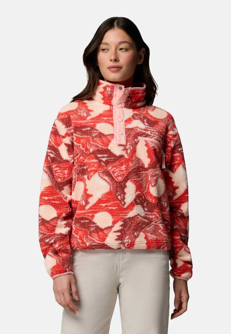 Femme portant un pull polaire à motifs rouges, roses et crème avec un col haut et un pantalon beige, tournée légèrement vers la gauche, les mains le long du corps.