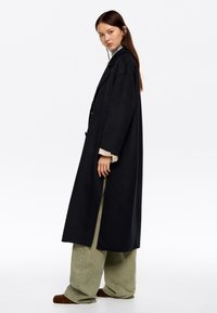 Mujer vistiendo un abrigo largo negro, pantalones de pierna ancha verde claro y zapatos marrones, de pie contra un fondo blanco liso.