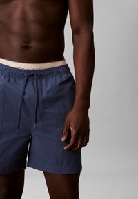 Pantaloni da bagno blu navy con cintura elastica e cordino. Presentano una leggera texture del tessuto e tasche laterali. Logo del marchio visibile sulla cintura.