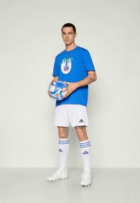 Zils kokvilnas T-krekls ar Itālijas grafiku, apvienots ar baltiem šortiem, baltiem futbola zeķēm ar zilām svītrām un baltiem sporta apaviem, turot futbola bumbu.