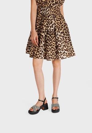 Donna che indossa una gonna a balze con stampa leopardata e sandali con cinturino intrecciato bianco e nero con tacco a blocco, in piedi contro uno sfondo semplice.