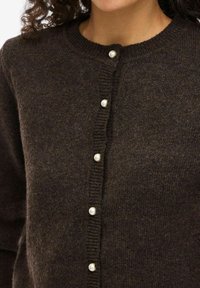 Mørkebrun strikket cardigan med perleaktige knapper nedover fronten og en rund ribbet hals på en person.