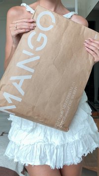 Brown paper shopping bag with white "MANGO" logo, wrinkled texture, holding a white frilly skirt, displayed against a neutral background.