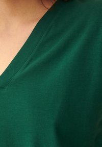 V-paita vihreänä, valmistettu sileästä puuvillasta ja lyhyillä hihoilla. Kangas on pehmeä tekstuuriltaan, ja kaula-aukossa on siisti, ommeltu reuna.