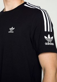 Camiseta de algodón negra con diseño de tres rayas blancas en los hombros, con pequeño logo blanco de Adidas en el pecho y la manga.