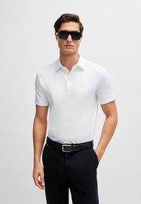 White polo shirt with short sleeves, flat collar, and subtle logo, paired with black tailored pants and a black belt. Dark sunglasses complete the look.