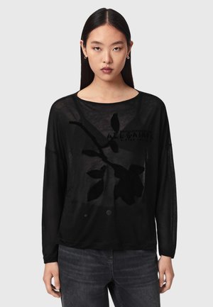 Kvinde med langt glat hår iført en sort gennemsigtig langærmet top med blomsterprint og mørke jeans mod en ensfarvet baggrund.