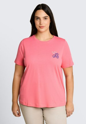 Femme aux longs cheveux foncés portant un t-shirt rose corail avec un petit motif scooter bleu et un texte sur le côté gauche de la poitrine.