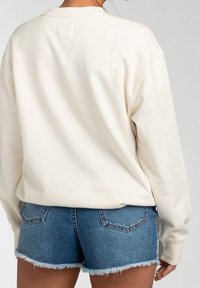 Osoba v jednoduché bílé mikině s dlouhými rukávy a modrých džínových krátkých šortkách s třepenými okraji, otočená zády k fotoaparátu.