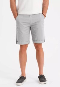 Shorts gris clair avec ourlets retroussés, fabriqués en tissu lisse. Le mannequin se tient avec les mains dans les poches, assortis à des chaussures foncées.