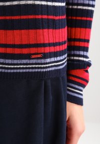 Suéter de punto con rayas horizontales rojas, azul marino y púrpuras. Presenta puños y dobladillo acanalados, con una pequeña etiqueta de marca cerca de la parte inferior.