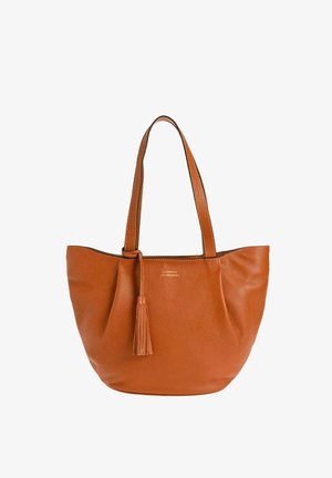 Sac cabas en cuir marron avec deux anses, fond incurvé et un détail pompon suspendu sur une anse.