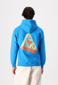 Blue hoodie featuring a geometric triangle design on the back, with pastel colors and "HUF" lettering. Soft fabric, relaxed fit.