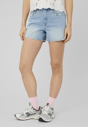 Short en denim avec broderie florale rose porté avec un haut blanc ajouré, des chaussettes roses et des chaussures de sport argentées sur une personne debout.