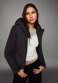 Giacca imbottita nera con cappuccio, caratterizzata da sezioni trapuntate, chiusura con zip e polsini a coste. Indossata sopra una canotta grigio chiaro.