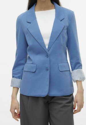 Femme portant un blazer bleu clair avec des poignets rayés retroussés, un haut blanc et un pantalon gris foncé, debout sur un fond blanc.