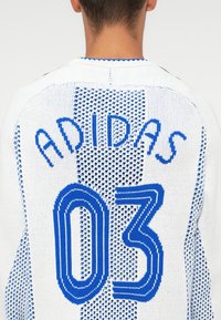 Bel bela pletena majica z modrim napisom "ADIDAS" in številko "03" v poudarjenem, plastehnem dizajnu. Geometrijski vzorec po celotni tkanini.