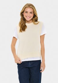 Glimlachende vrouw met lang blond haar wearing een crèmekleurige mouwloze gebreide vest over een wit t-shirt en donkerblauwe jeans tegen een effen achtergrond.