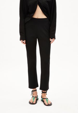 Osoba ubrana w czarne spodnie o prostych nogawkach, czarną koszulę z długim rękawem częściowo rozpiętą oraz czarne sandały z zielonymi i czarnymi paskami.