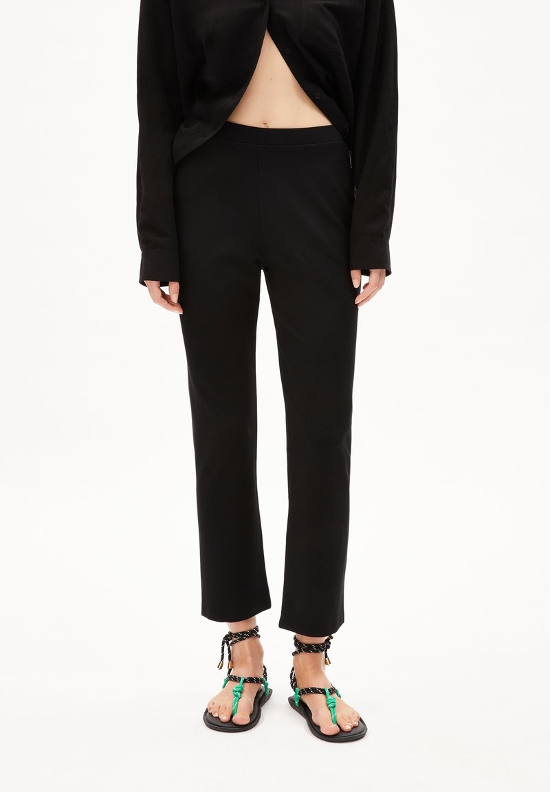 Personne portant un pantalon noir à jambes droites, une chemise noire à manches longues partiellement boutonnée, et des sandales noires avec des brides vertes et noires.