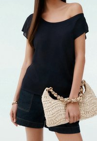 Zwarte off-shoulder top van zachte stof; gecombineerd met donkere denim shorts. Houdt een geweven tas met een gouden kettinghandvat vast.