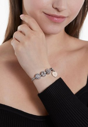Silver charm bracelet with decorative beads, featuring floral designs and a heart-shaped tag. Worn on a wrist with a black top.