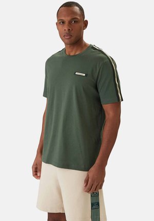 Uomo che indossa una maglietta verde scuro a maniche corte con nastro a righe sulle spalle e pantaloncini beige con una toppa verticale con logo sulla gamba.