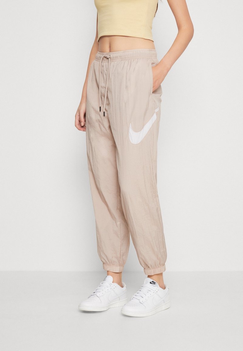 Beige Nike joggers with an elastic waistband, side pockets, and a white swoosh logo. The fabric has a smooth, lightweight texture.