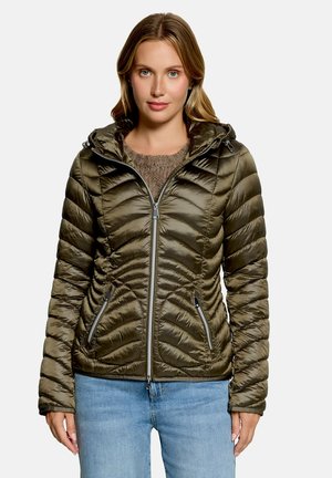 Frau mit blondem Haar, die eine olivgrüne gesteppte Jacke mit Kapuze, hellblaue Jeans und einen braunen Pullover darunter trägt, nach vorne blickend.