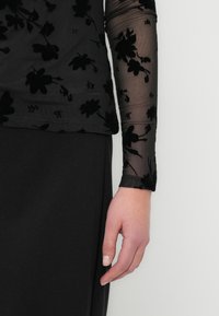 Μαύρο τοπ με φλοράλ μοτίβο, κατασκευασμένο από διάφανο ύφασμα, με μακριά μανίκια και υφή σχεδίου. Το ρούχο συνδυάζεται με μια μονόχρωμη μαύρη φούστα.