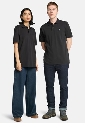 Polo shirt nera a maniche corte, accompagnata da pantaloni di denim blu larghi a sinistra e jeans scuri slim con stivali verdi a destra.