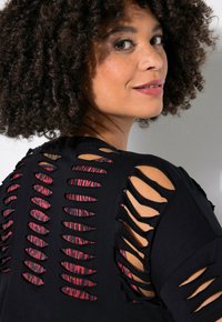 Černý top s výřezovými detaily, odhalující vrstvení s červeným a černým vzorem uvnitř, má krátké rukávy a volný střih.