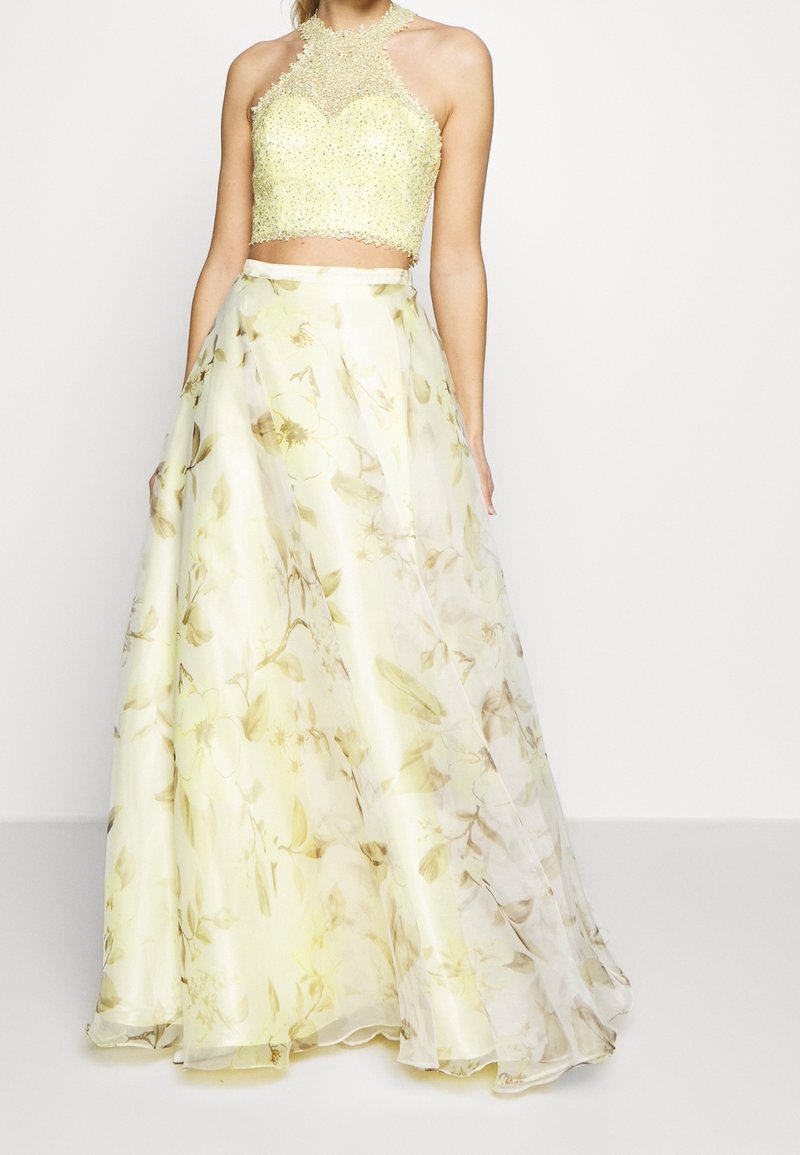 Femme portant une robe formelle deux pièces avec un haut dos nu orné de perles et une longue jupe fluide à motif floral dans des tons jaunes doux.