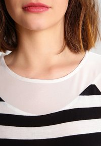 Svart- och vitrandig topp med en luftig, genomskinlig halsringning. Kortärmad och mjuk i texturen; har en rund, tight krage.