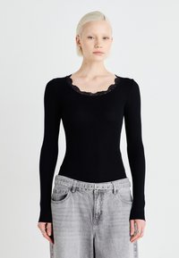 Top preto de manga longa com textura canelada e decote em renda com bordas onduladas; design ajustado. Modelo combinado com jeans cinzentos de cintura alta. Acessórios simples.