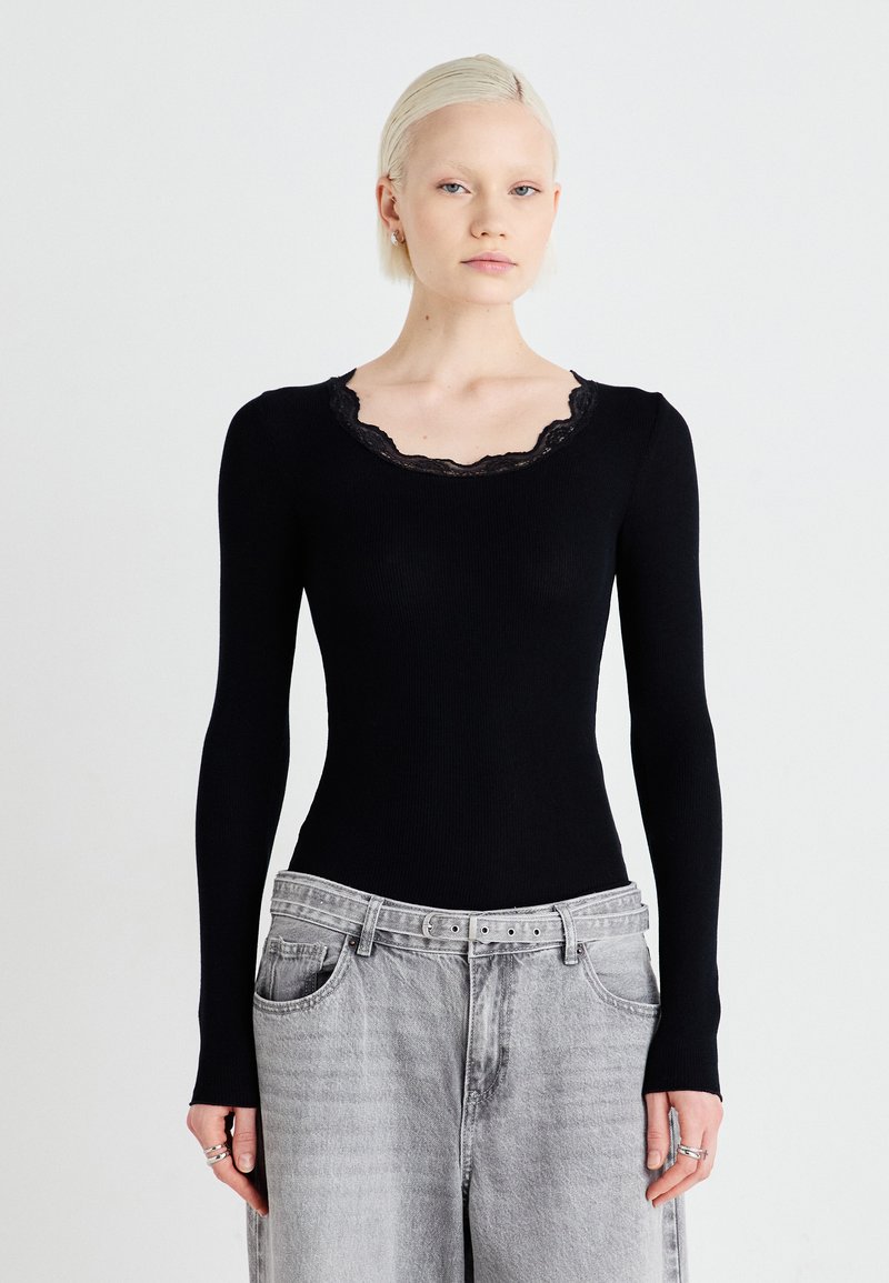 Top preto de manga longa com textura canelada e decote em renda com bordas onduladas; design ajustado. Modelo combinado com jeans cinzentos de cintura alta. Acessórios simples.