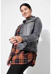 Harmaa farkkutakki rintataskuilla oranssi-mustan ruudullisen paidan päällä. Mallilla on päällään mustat legginsit ja kerroksittain sormuksia; tausta on yksinkertainen valkoinen.