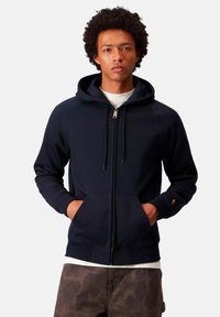 Un giovane uomo indossa una felpa blu navy con zip e tasche, una maglietta bianca sotto e pantaloni mimetici marroni, in piedi contro uno sfondo chiaro e semplicemente.