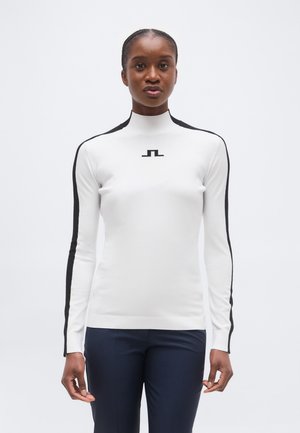 Naist, kes kannab valget, pikkade varrukatega särk mustade triipudega varrukatel ja väikese musta logoga rinnal, ning tumedaid pükse.